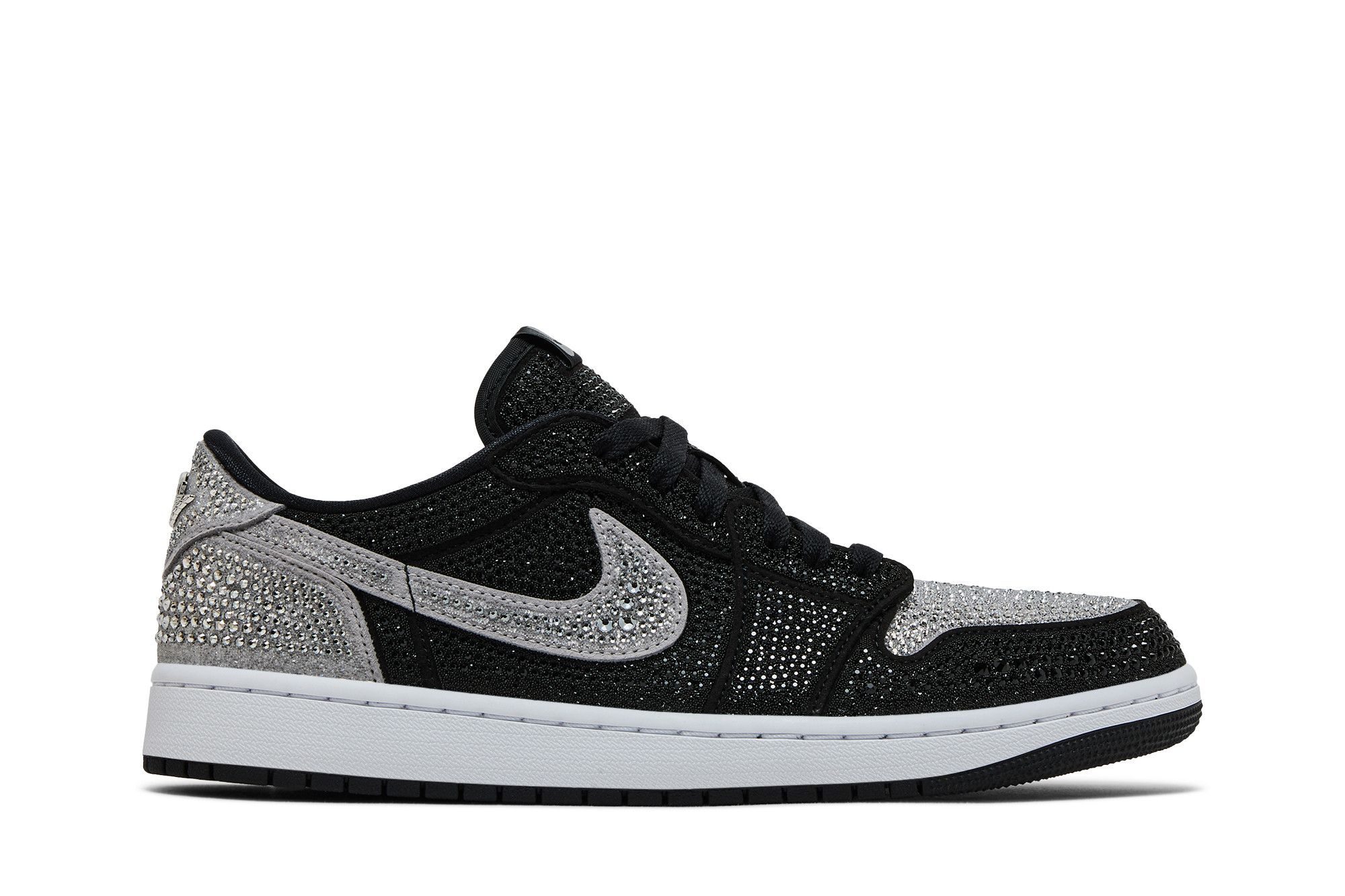 Imagem lateral esquerda de um tênis feminino Swarovski x Air Jordan 1 Retro Low OG 'Shadow' exibindo um fundo branco. O tênis apresenta uma combinação de cores preta e cinza, com detalhes brilhantes de cristais Swarovski cobrindo partes da superfície, incluindo o swoosh da Nike, o calcanhar e o bico. O design é detalhado e elegante, focado na exclusividade da colaboração entre Swarovski e Air Jord.