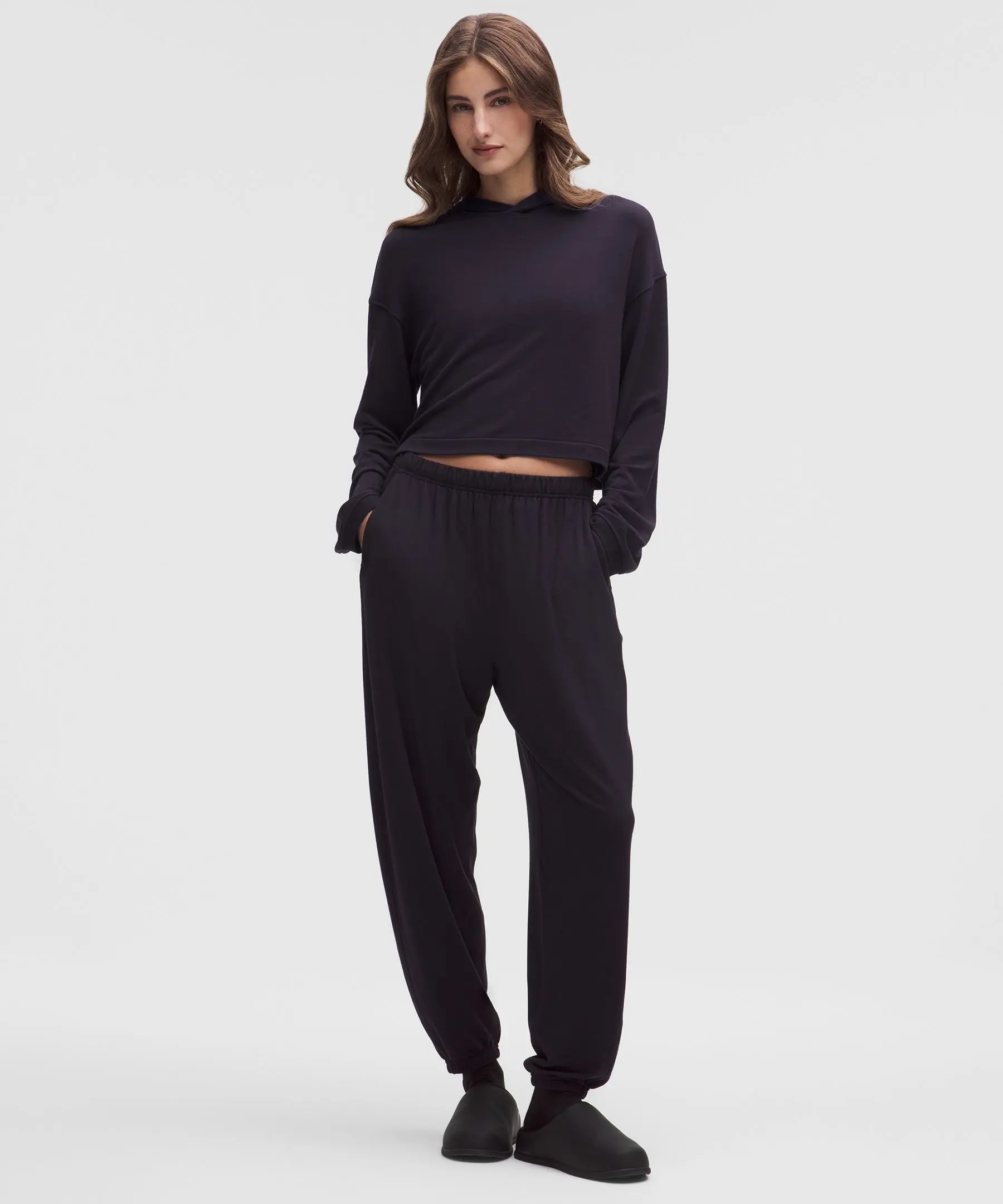 Imagem de corpo inteiro de mulher usando conjunto preto BeCalm, com moletom cropped e calça esportiva. Ela está de pé com as mãos nos bolsos e olhar leve para baixo, fundo branco.