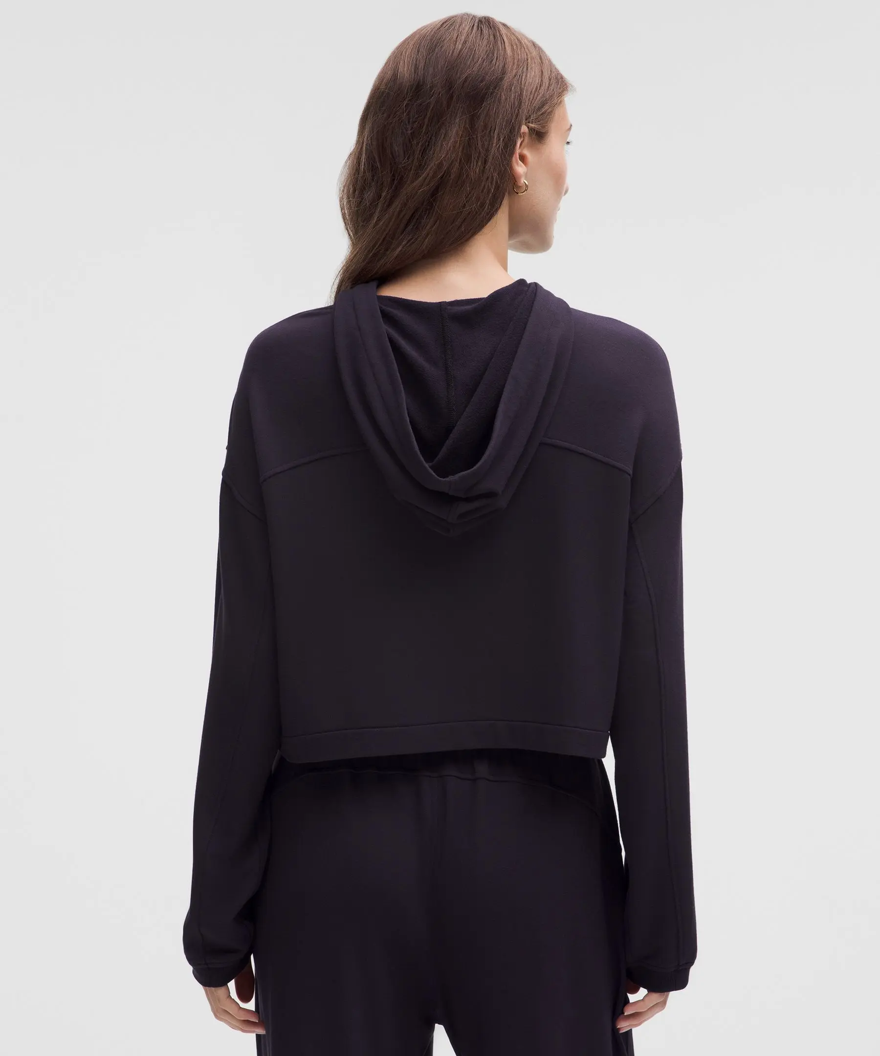 Vista traseira de mulher usando moletom preto BeCalm cropped com capuz caído, mostrando detalhes da costura. Fundo branco simples e iluminação uniforme.